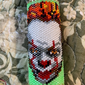 May include: A beaded lighter case with a pixelated clown face design. The case is mainly white, with red, grey, and brown accents, and a green base. The top section has orange, brown, and yellow beads. The lighter is silver.