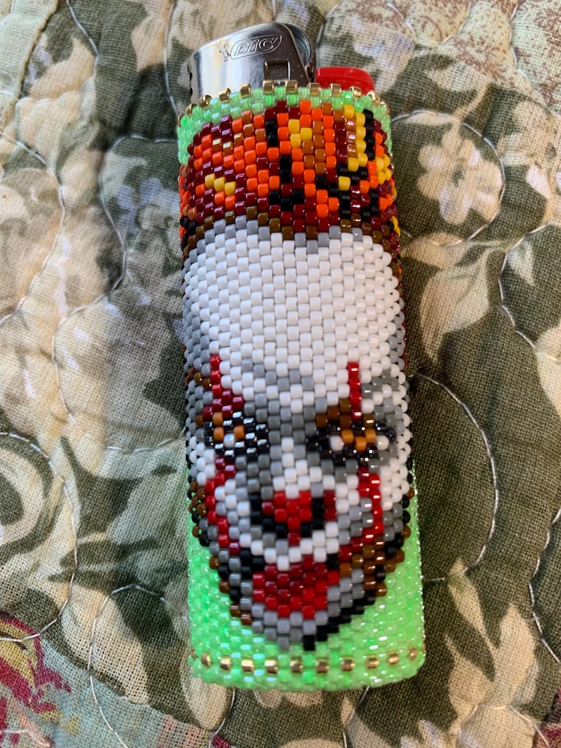 May include: A beaded lighter case with a pixelated clown face design. The case is mainly white, with red, grey, and brown accents, and a green base. The top section has orange, brown, and yellow beads. The lighter is silver.