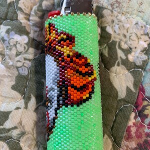 May include: A green beaded lighter case featuring a butterfly design. The butterfly is detailed with orange, yellow, black, and white beads. Gold-coloured beads accent the top and bottom of the case.