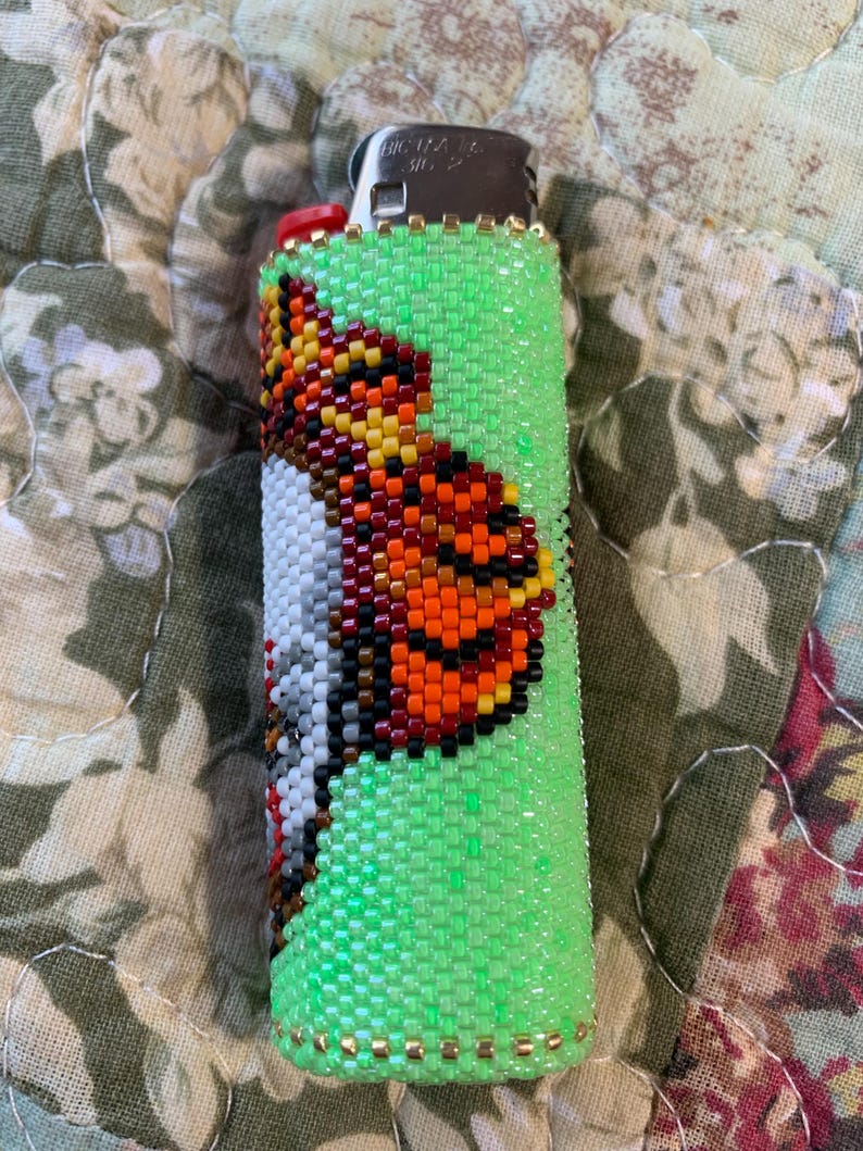 May include: A green beaded lighter case featuring a butterfly design. The butterfly is detailed with orange, yellow, black, and white beads. Gold-coloured beads accent the top and bottom of the case.