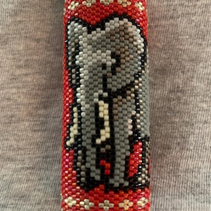 May include: A rectangular lighter encased in a beaded sleeve featuring a grey elephant design. The background of the sleeve is composed of small red beads with a geometric pattern of silver beads at the top and bottom edges of the lighter.