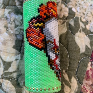 May include: A green beaded lighter cover featuring a butterfly design. The butterfly is crafted from orange, black, white, and grey beads. The lighter cover is embellished with gold-coloured beads.