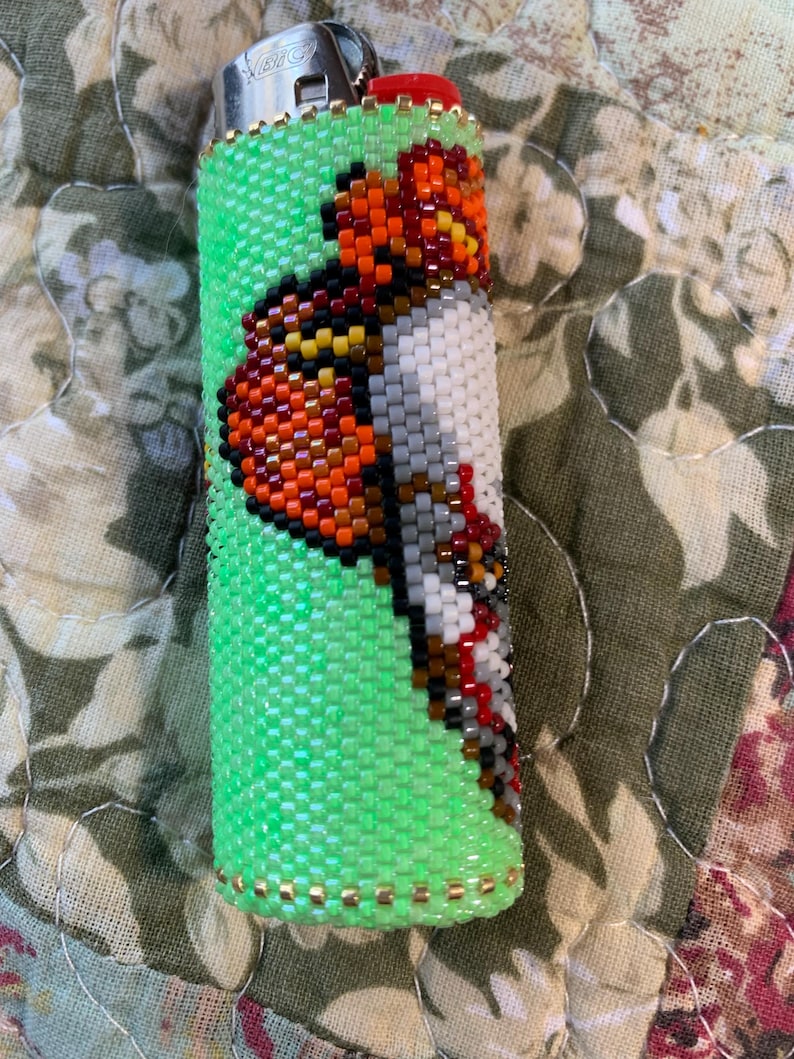 May include: A green beaded lighter cover featuring a butterfly design. The butterfly is crafted from orange, black, white, and grey beads. The lighter cover is embellished with gold-coloured beads.