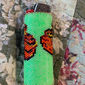 May include: A green beaded lighter cover featuring a butterfly design in orange, black, and yellow. The cover is made of small, evenly spaced beads and has a gold trim. The lighter has a red button. The cover is a decorative accessory.