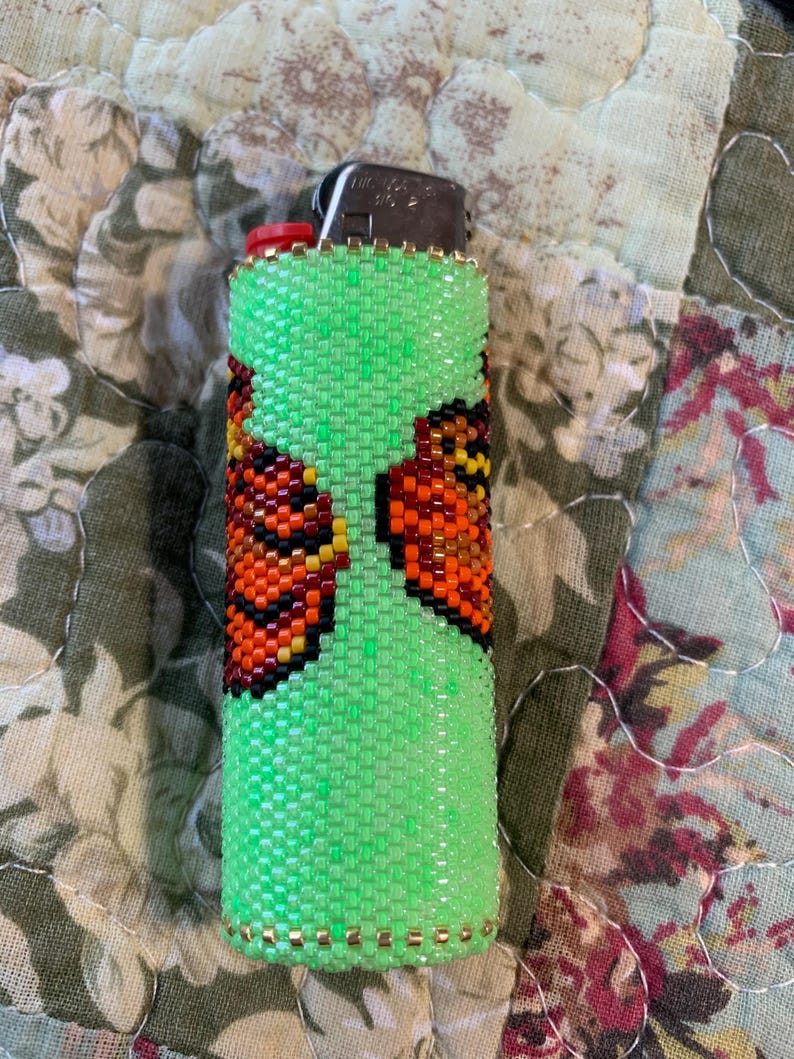 May include: A green beaded lighter cover featuring a butterfly design in orange, black, and yellow. The cover is made of small, evenly spaced beads and has a gold trim. The lighter has a red button. The cover is a decorative accessory.