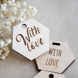 Puede incluir: Tres etiquetas hexagonales de madera con el texto "With Love" grabado en ellas. Las etiquetas son de madera marrón claro y tienen un agujero en la parte superior para colgarlas.