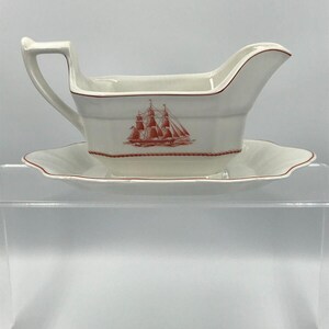 May include: White porcelain gravy boat with a red ship design and a matching saucer. The boat has a handle and a wide, shallow bowl. The saucer has a scalloped edge.