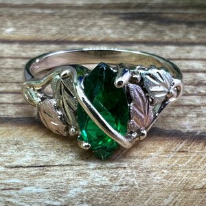 May include: A silver ring featuring a green gemstone set within gold and silver leaves.