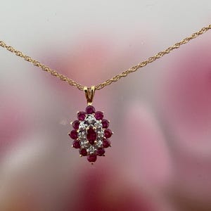 Op de afbeelding: Een gouden ketting met een ovaalvormige hanger. De hanger is bezet met een centrale robijnrode edelsteen in marquise-slijpvorm, omringd door een halo van kleine witte diamanten en een buitenste ring van ronde robijnrode edelstenen.