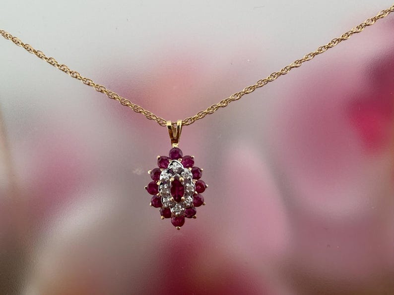 Op de afbeelding: Een gouden ketting met een ovaalvormige hanger. De hanger is bezet met een centrale robijnrode edelsteen in marquise-slijpvorm, omringd door een halo van kleine witte diamanten en een buitenste ring van ronde robijnrode edelstenen.