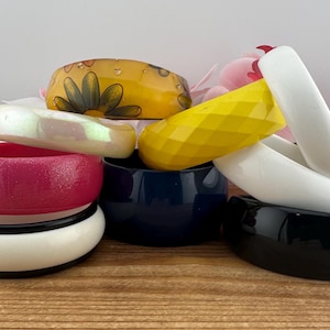 May include: A selection of colourful bangles in different styles. Features a yellow bangle with a flower pattern, a faceted yellow bangle, and white, pink, and black bangles. The bangles are stacked on a wooden surface.