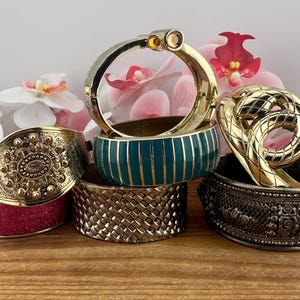 May include: A collection of ornate bangles in various styles and colors. The bangles include gold, silver, and teal designs, some with intricate patterns and embellishments. A pink glitter bangle is also present.