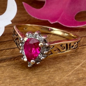 May include: A gold ring featuring a pear-shaped red gemstone at the centre, surrounded by a halo of small clear stones. The band is crafted with an intricate openwork filigree design, providing a detailed and ornate appearance to the metalwork.