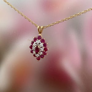 Op de afbeelding: Een gouden ketting met een hanger voorzien van een centrale robijn in marquise-slijpvorm, omringd door een halo van kleine ronde diamanten, die verder is omsloten door een cluster van ronde robijnen in een bloemvormige zetting.