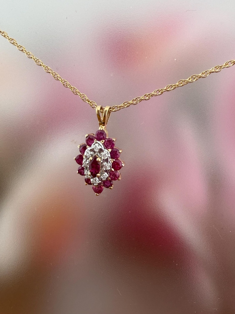 Op de afbeelding: Een gouden ketting met een hanger voorzien van een centrale robijn in marquise-slijpvorm, omringd door een halo van kleine ronde diamanten, die verder is omsloten door een cluster van ronde robijnen in een bloemvormige zetting.