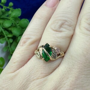May include: A gold ring featuring a green gemstone set within a leaf design. The ring is worn on a finger.