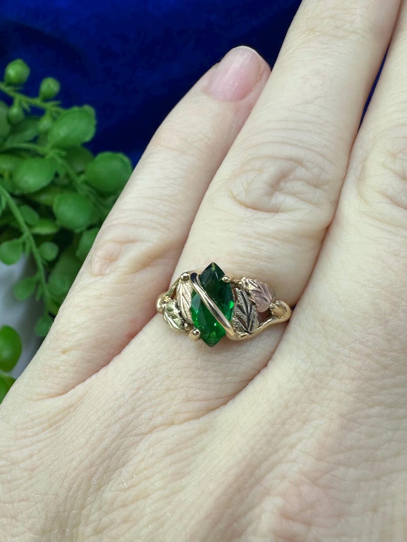May include: A gold ring featuring a green gemstone set within a leaf design. The ring is worn on a finger.