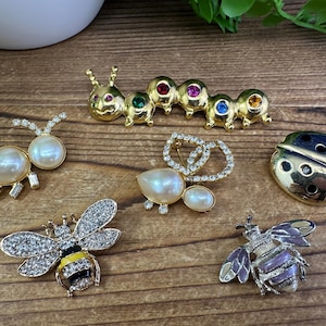 Peut inclure: Une collection de neuf broches dorées présentant différents motifs, dont une chenille avec des pierres précieuses colorées, une coccinelle avec des accents noirs et dorés, une abeille avec des accents noirs et jaunes et plusieurs broches en forme de perle.