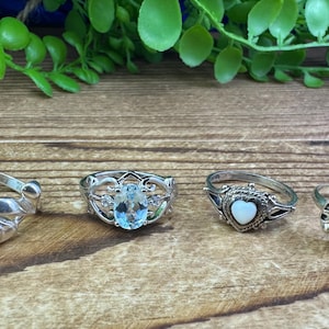 May include: Four silver rings with different gemstones. The rings feature a dolphin, a heart-shaped stone, a blue oval stone, and a red round stone.