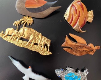 May include: A collection of six wooden and metal animal brooches. The brooches include a duck, a fish, a bird, a group of elephants, a seagull, and a frog. The frog brooch is decorated with turquoise stones.