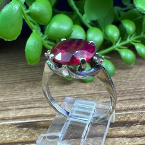 May include: A silver ring with a large, oval-cut, deep red gemstone. The gemstone is held in place by prongs, and the band has a simple design. The ring is displayed on a clear plastic stand, with green foliage in the background.