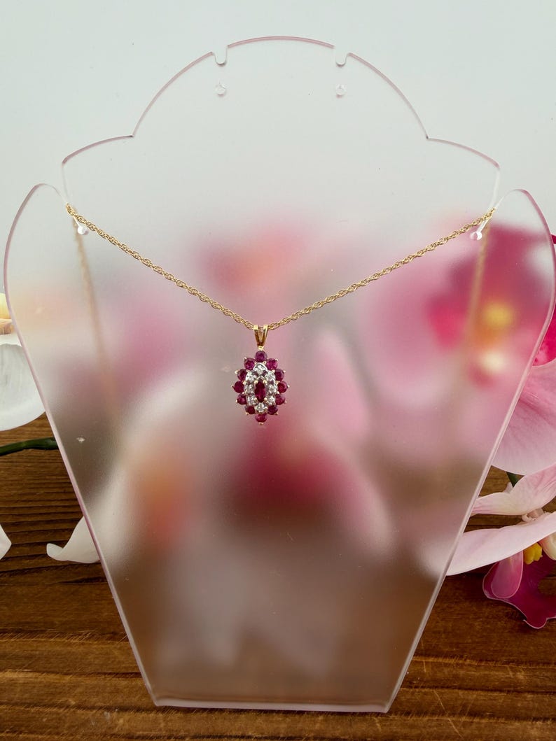 Op de afbeelding: Een gouden touwketting met een marquisevormige hanger. De hanger is bezet met een centrale robijn in marquiseslijpsel, omringd door een halo van kleine ronde witte diamanten en een buitenrand van ronde robijnen in een gouden zetting.