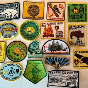 May include: A collection of 15 colourful embroidered patches featuring the text "Klondike Derby" and various years from 1988 to 2024. The patches are yellow, blue, green, and white with black and red embroidery.