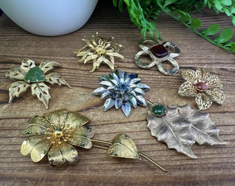 Peut inclure: Un ensemble de sept broches dorées représentant différents motifs floraux et de feuilles. Les broches sont ornées de pierres colorées, notamment vertes, rouges et bleues.