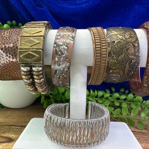 May include: A set of eight metal bracelets in various styles and finishes, including hammered copper, brass, and silver. The bracelets are displayed on a white stand.