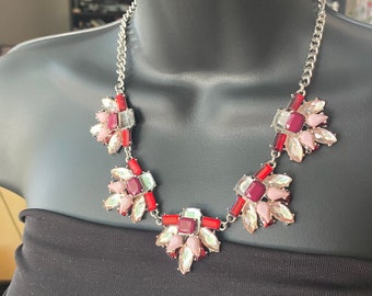 May include: A silver chain necklace with a statement pendant featuring a cluster of red, pink, and clear faceted gemstones. The pendant is shaped like a flower with multiple points.