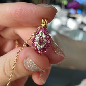 Op de afbeelding: Een gouden hanger met een centrale marquise-geslepen rode edelsteen, omringd door een halo van kleine witte steentjes en een buitenrand van ronde rode edelstenen, bevestigd aan een fijne gouden koordketting.