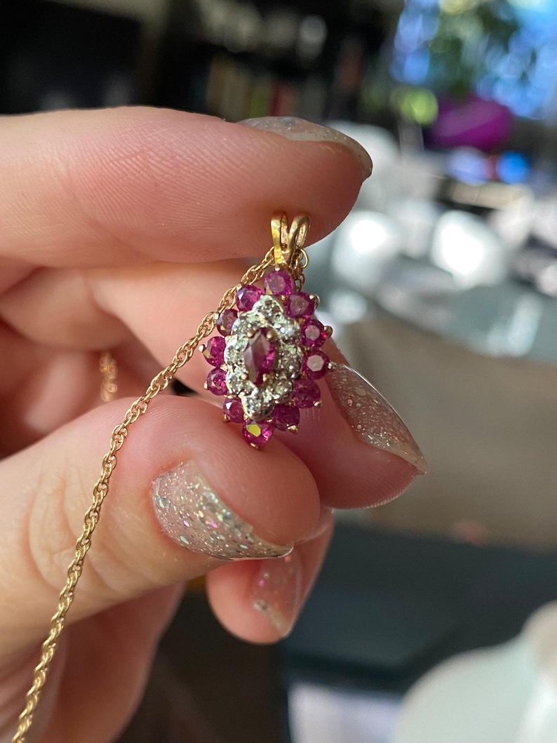 Op de afbeelding: Een gouden hanger met een centrale marquise-geslepen rode edelsteen, omringd door een halo van kleine witte steentjes en een buitenrand van ronde rode edelstenen, bevestigd aan een fijne gouden koordketting.