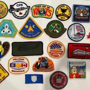 May include: A collection of colourful embroidered patches featuring various designs, including a beaver, a helicopter, a scout's emblem, and a cartoon character. The patches are arranged on a white surface.