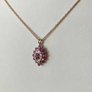Op de afbeelding: Een gouden ketting met een hanger voorzien van een centrale donkerroze edelsteen in marquise-slijpvorm, omringd door een halo van kleine witte steentjes en een buitenrand van ronde donkerroze edelstenen, gezet in een goudkleurig metalen frame.