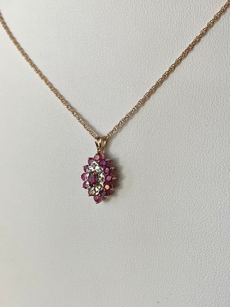 Op de afbeelding: Een gouden ketting met een hanger voorzien van een centrale donkerroze edelsteen in marquise-slijpvorm, omringd door een halo van kleine witte steentjes en een buitenrand van ronde donkerroze edelstenen, gezet in een goudkleurig metalen frame.