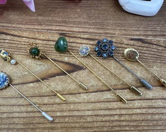 May include: Seven gold-toned hat pins with various decorative tops. The tops include a green stone, a blue flower, a gold flower, a white pearl, a blue and white floral design, and a gold design.