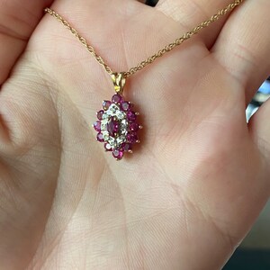 Op de afbeelding: Een gouden ketting met een hanger voorzien van een centrale robijn in marquise-slijpvorm, omringd door een halo van kleine ronde diamanten, die verder is omsloten door een cluster van ronde robijnen in een geelgouden zetting met oogje.
