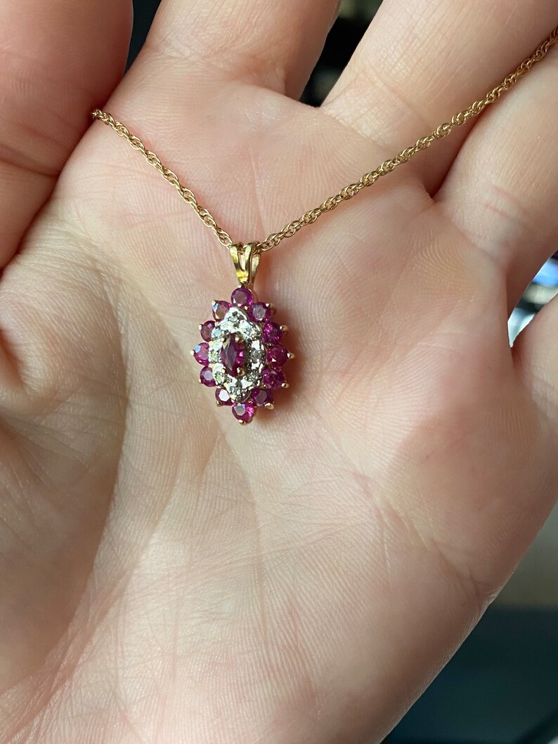 Op de afbeelding: Een gouden ketting met een hanger voorzien van een centrale robijn in marquise-slijpvorm, omringd door een halo van kleine ronde diamanten, die verder is omsloten door een cluster van ronde robijnen in een geelgouden zetting met oogje.