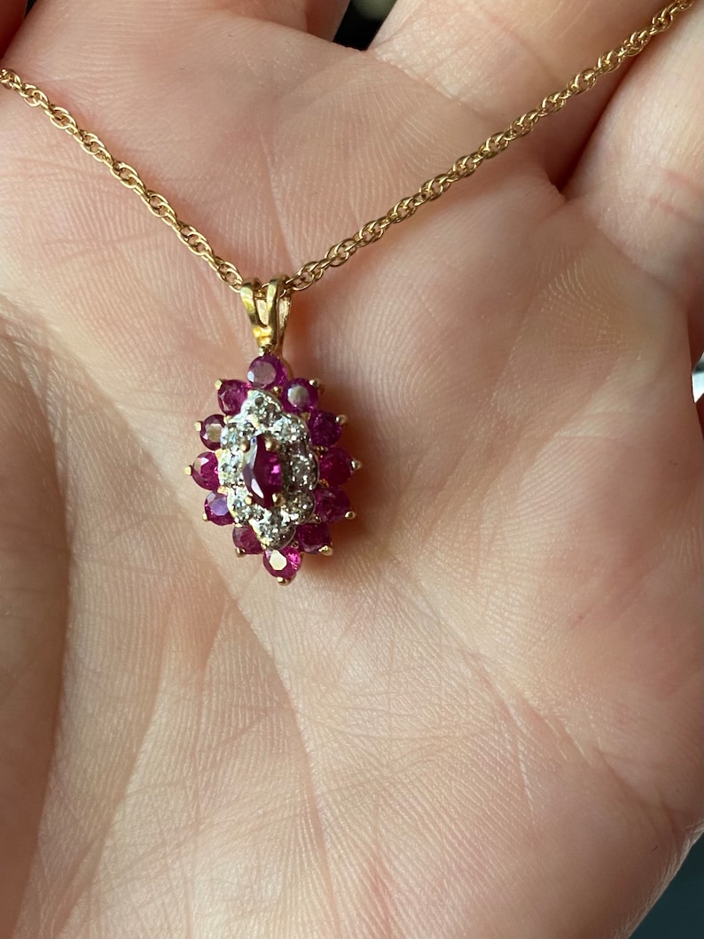 Op de afbeelding: Een gouden ketting met een marquisevormige hanger. De hanger is bezet met een centrale robijn in marquise-slijpvorm, omringd door een halo van kleine ronde diamanten, die weer is omringd door een cluster van ronde robijnen in gouden zettingen.