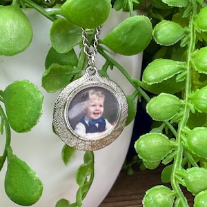 May include: Silver necklace with a round photo locket featuring a young child wearing a white shirt and a blue bow tie.