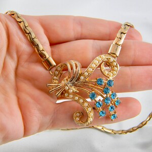 May include: A gold chain necklace with a pendant featuring a cluster of blue rhinestones and faux pearls in a floral design.