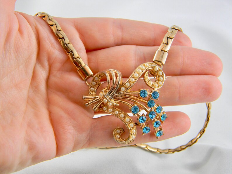 May include: A gold chain necklace with a pendant featuring a cluster of blue rhinestones and faux pearls in a floral design.