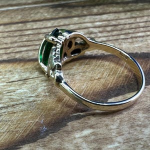 May include: A silver ring with a large, emerald green gemstone set in a filigree design. The ring is on a wooden surface.