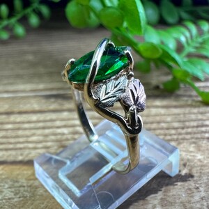 May include: A gold ring featuring a large green emerald-cut gemstone and a leaf design. The ring is displayed on a clear stand.
