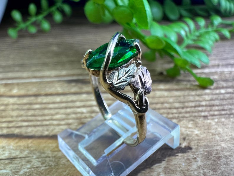 May include: A gold ring featuring a large green emerald-cut gemstone and a leaf design. The ring is displayed on a clear stand.