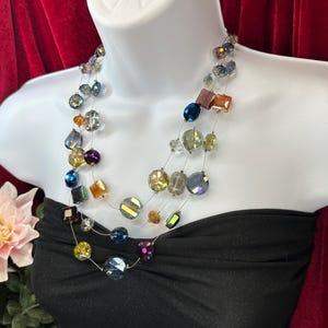 May include: A multi-strand necklace with iridescent beads in various shapes and colours, including blue, yellow, purple, and orange. The necklace is displayed on a white mannequin bust against a red backdrop. The necklace is a fashion accessory.