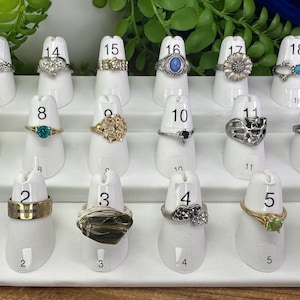 Peut inclure: Un présentoir de 18 bagues avec différentes pierres et motifs sur des supports à bagues blancs numérotés de 1 à 18. Les bagues sont disposées en trois rangées.