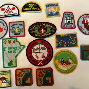 May include: A collection of 16 colourful embroidered scout badges. The badges feature various designs, including a squirrel, a maple leaf, a tent, a canoe, and a campfire. The badges are all in different colours and sizes, and they are all in good condition.