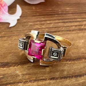 May include: A gold-toned ring with a large, square, magenta-colored gemstone at its center. The ring features silver-toned accents and two smaller square details on either side of the central stone. The ring is set against a wooden surface.