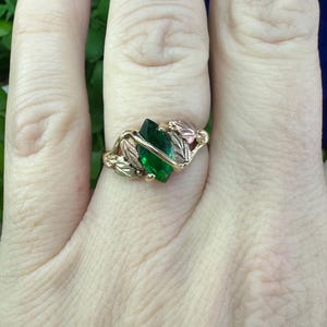 May include: A gold ring featuring a green gemstone and leaf accents. The ring is worn on a finger.